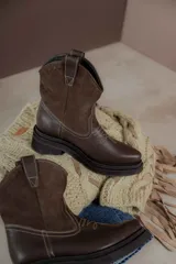 Bota estilo western de caña media, confeccionada en una combinación de cuero texturizado en la parte inferior y gamuza en la caña. Presenta costuras decorativas en contraste y suela de goma robusta con plataforma.