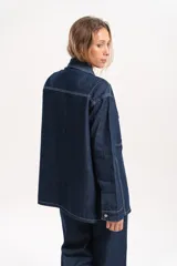 Sobrecamisa de jean oversize en color azul oscuro, con costuras a la vista en hilo blanco que resaltan su diseño. Presenta cuello clásico, cierre frontal con botones y bolsillos delanteros.