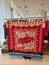 Pañuelo cuadrado de seda con estampado geométrico y floral en tonos rojos, rosas, amarillos y azules. Incluye la inscripción "Lamode" en el centro.