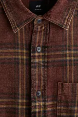 Camisa de franela de algodón suave con efecto lavado, color bordó con estampado a cuadros en tonos negros y amarillos. Presenta un corte relajado, cuello inglés, tapeta clásica de botones, un bolsillo superior abierto y canesú en la espalda. Tiene mangas largas con puños abotonados y un bajo ligeramente redondeado.