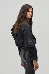 Camisa negra de popelina de algodón con cuello clásico y cierre frontal de botones de nácar. Presenta volados dobles que recorren el frente y la espalda desde el hombro hasta la cintura, y volados adicionales en las mangas a la altura de los codos.