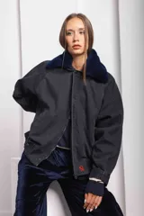 Campera oversize de jean color negro con cuello de corderito azul, cierre frontal con cremallera y solapa con botones a presión. Presenta mangas amplias y un pequeño bordado rojo en el puño.