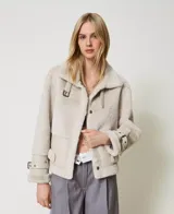 Chaqueta color beige de napa y piel sintética con cuello con cierre de hebilla y almeja, mangas largas con hombros caídos, tirantes en los puños, cierre de cremallera oculta, bolsillos laterales, cierre de hebilla y almeja en el dobladillo y placa en T ovalada en la parte posterior.