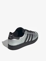 Championes Adidas Superstar con acabado metalizado plateado, caracterizadas por su icónica puntera de goma y las tres bandas laterales en color negro. Presentan una suela de plataforma negra y detalles en contraste en el talón y la lengüeta.