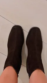Bota de caña alta de gamuza en color marrón chocolate, con taco cuadrado y puntera cuadrada. Cuenta con un diseño holgado en la caña y detalle de trabilla decorativa en el tobillo.