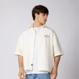 Camisa de manga corta color marfil, corte boxy, con cuello camisero, cierre frontal de botones y pequeño bordado marrón en el pecho con las siglas VCP.