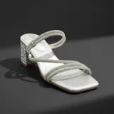 Sandalia de tacón cuadrado medio, color plateado, con tiras finas recubiertas de strass. Presenta puntera cuadrada y una pulsera ajustable al tobillo que puede retirarse para usar como chinela.