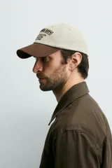Gorra de seis paneles de algodón en color beige con visera marrón contrastante. Cuenta con un diseño bordado en marrón en el frente con texto alusivo.