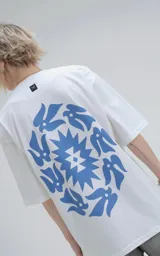 Remera blanca de corte holgado con estampa central en color azul que combina motivos geométricos tipo chakana con figuras de aves dispuestas en círculo.