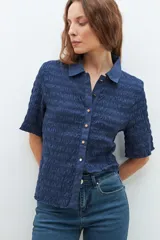 Camisa de manga corta en color bordó, confeccionada en tejido con textura de punto smock. Presenta cuello clásico, cierre frontal con botones y un ajuste entallado al cuerpo.
