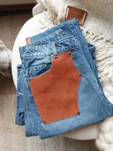Pantalón de jean celeste con cintura elastizada, corte recto, bolsillos laterales y aplique de cuero marrón en el frente. Presenta botones metálicos en los laterales y etiqueta de cuero en la parte posterior.
