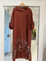 Vestido largo terracota de lino, con diseño camisero, botones frontales, bolsillos con solapa estampada en el pecho y detalle de estampado floral en el ruedo. Mangas largas con presillas para arremangar.