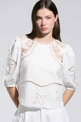 Top Allegra de color blanco, con mangas abullonadas a la altura del codo y puños elásticos. Presenta detalles de calados y bordados florales en los hombros, mangas y ruedo.