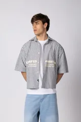Camisa de manga corta boxy fit con estampado de rayas verticales en tonos gris y blanco, con detalles de texto estampado en la parte inferior frontal y botones personalizados.