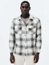 Camisa de manga larga a cuadros blancos, grises y negros, con cuello clásico, cierre frontal con botones, dos bolsillos con solapa abotonada en el pecho y puños abotonados.