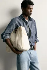 Bolso tipo shopper de tejido color crudo, con doble asa de hombro y detalles en cuero negro. Cuenta con compartimento principal con cierre de tira y botón a presión.