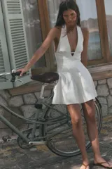 Vestido mini de color blanco con cuello halter, escote en pico y falda con vuelo tipo capa. Presenta cierre con cremallera oculta en la espalda y lazada en el cuello.