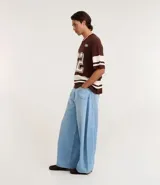 Remera oversized deportiva color marrón con cuello en V, rayas blancas en las mangas y en la parte inferior, y el número "62" estampado en blanco en el frente. Incluye el bordado "Humanity" y un logo de globo terráqueo.