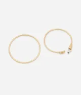 Brazaletes rígidos dorados en kit de dos unidades. Incluye un brazalete cerrado de diseño liso y un brazalete abierto tipo cuff con terminaciones decorativas en forma de hoja y un cristal artificial.