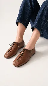 Zapato de cuero color chocolate con cordones finos negros, puntera cuadrada y detalles de perforaciones decorativas en los laterales.