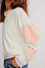 Buzo liviano de algodón, de calce amplio y color crudo, con escote redondo, hombros caídos y aplicaciones de corazones rosados en las mangas. Cuenta con detalles de cinta en contraste en el cuello, los puños y el ruedo.