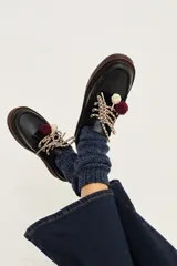Zapato tipo mocasín de cuero negro con cordones trenzados, detalle de pompones bordó y nudos decorativos. Presenta suela de goma con borde en contraste.