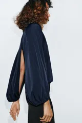 Top azul marino de tejido fluido con cuello redondo y manga larga abullonada con aberturas. Presenta una abertura en la espalda con tira del mismo tejido y cierre de ganchos metálicos.