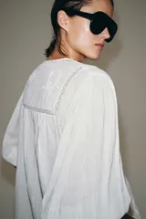 Camisa blanca de algodón con escote en pico, mangas por debajo del codo y detalles de bordados y encaje a tono en el pecho y las mangas.