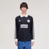 Camiseta Adidas de manga larga en colaboración con la banda Oasis, de color negro con cuello polo, puños y detalles en blanco marfil. Presenta las tres tiras características en las mangas, el logo trébol de Adidas, un escudo circular conmemorativo de la gira 'Live '25' y el estampado con la inscripción 'oasis' en el centro del pecho.