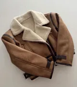 Chaqueta tipo gamulán de cuero sintético en color tostado, con interior y cuello de corderito sintético en color crema. Presenta cierre frontal cruzado, detalles de ribetes en cuero oscuro y correas ajustables con hebillas en los puños y laterales.
