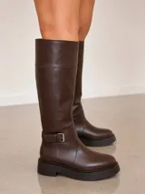 Bota de caña alta confeccionada en cuero negro, con plataforma y taco cuadrado. Presenta una tira decorativa con hebilla metálica en la zona del tobillo y un diseño de caña recta con costuras visibles.
