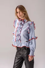 Camisa celeste con volados decorativos en los hombros, pecho, mangas y ruedo, destacados con ribetes en tono rojo. Presenta cierre frontal de botones, cuello alto y mangas largas abullonadas con puños ajustados.