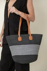Bolso tipo tote de paja color negro con rayas horizontales blancas y asas de cuero color marrón.