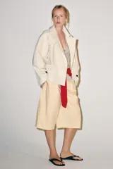 Trench corto color marfil de algodón, con cuello camisero, solapa cortaventos, mangas largas amplias con puños abotonados y cierre cruzado con un botón visible.