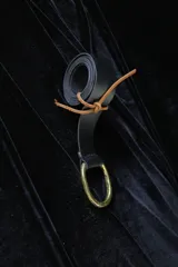 Cinturón de cuero negro enrollado, con hebilla metálica ovalada dorada y detalle de atado con tira de cuero marrón claro.
