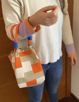 Cartera tipo bucket con diseño de bloques de color en tonos naranja, beige y crema. Confeccionada en lona resistente, cuenta con una manija de cuerda náutica naranja con detalles en azul y herrajes metálicos plateados.