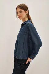 Camisa de denim azul oscuro con cuello clásico, cierre frontal de botones y mangas largas. Presenta un delicado bordado de motivos florales y vegetales realizado con cuentas y canutillos en tono azul marino.