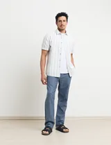 Camisa de manga corta a rayas verticales color natural y celeste, con cuello clásico, cierre frontal con botones de madera y bolsillo de parche en el pecho.