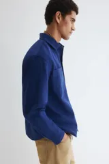 Sobrecamisa azul de lona gruesa de algodón, con cuello, tapeta con botones a presión y canesú en la espalda. Tiene mangas largas con botón a presión en los puños y dos bolsillos superiores de parche. El bajo es levemente redondeado y el corte es oversized.