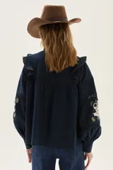 Blusa azul marino de algodón con volados en los hombros, mangas largas abullonadas y detalles de bordados florales y geométricos en el pecho y las mangas.