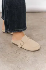 Zueco de cuero gamuzado color beige, con punta cerrada y costuras decorativas. Presenta una tira lateral con hebilla metálica dorada ajustable.