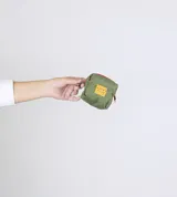 Monedero pequeño de tela color verde oliva, con cierre superior y etiqueta rectangular amarilla con logo de la marca en el frente. Incluye un pequeño gancho metálico lateral.