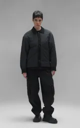 Campera tipo sobrecamisa de diseño oversize, confeccionada en microfibra opaca con patrón matelaseado geométrico. Presenta cuello, puños, cartera central y ruedo en gabardina de algodón, con cierre frontal mediante broches a presión. El diseño del ruedo simula una prenda superpuesta.