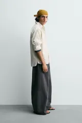 Camisa blanca de algodón con corte relajado, cuello solapa, manga larga con puño abotonado y bolsillo de plastrón en el pecho. Presenta bajo asimétrico con aberturas laterales y cierre frontal de botonadura.