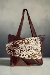 Cartera de cuero marrón estilo tote bag, con diseño en patchwork y bolsillo frontal de pelo con estampado animal print tipo vaca. Cuenta con doble asa de hombro y detalles metálicos.
