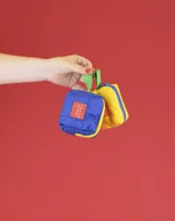 Monedero pequeño de tela color beige con cierre azul y tira de agarre color verde. Incluye un parche rectangular rojo con logo y gancho metálico para colgar.