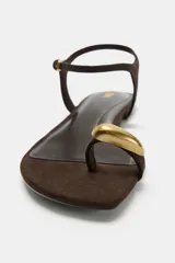 Sandalia plana de color marrón con diseño de dedo, tira trasera ajustable con hebilla metálica y detalle de adorno metálico dorado en la parte frontal.