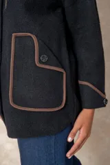 Chaqueta negra de corte amplio y diseño estructurado, con cierre frontal de botones. Presenta bolsillos frontales y puños con ribetes en contraste color marrón.