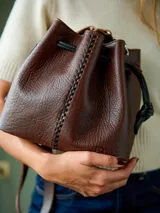 Cartera tipo bucket de cuero graneado color chocolate, con detalle de costura central con tientos a contratono en color negro. Cuenta con cierre de ajuste mediante tiras, correa regulable y diseño de formato clásico con aire bohemio.