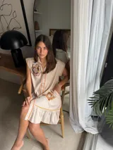 Vestido corto beige de algodón con cuello camisero y cierre frontal de botones. Presenta mangas cortas y detalles bordados a contratono en la zona del pecho y en los bolsillos, además de un volado en el ruedo.